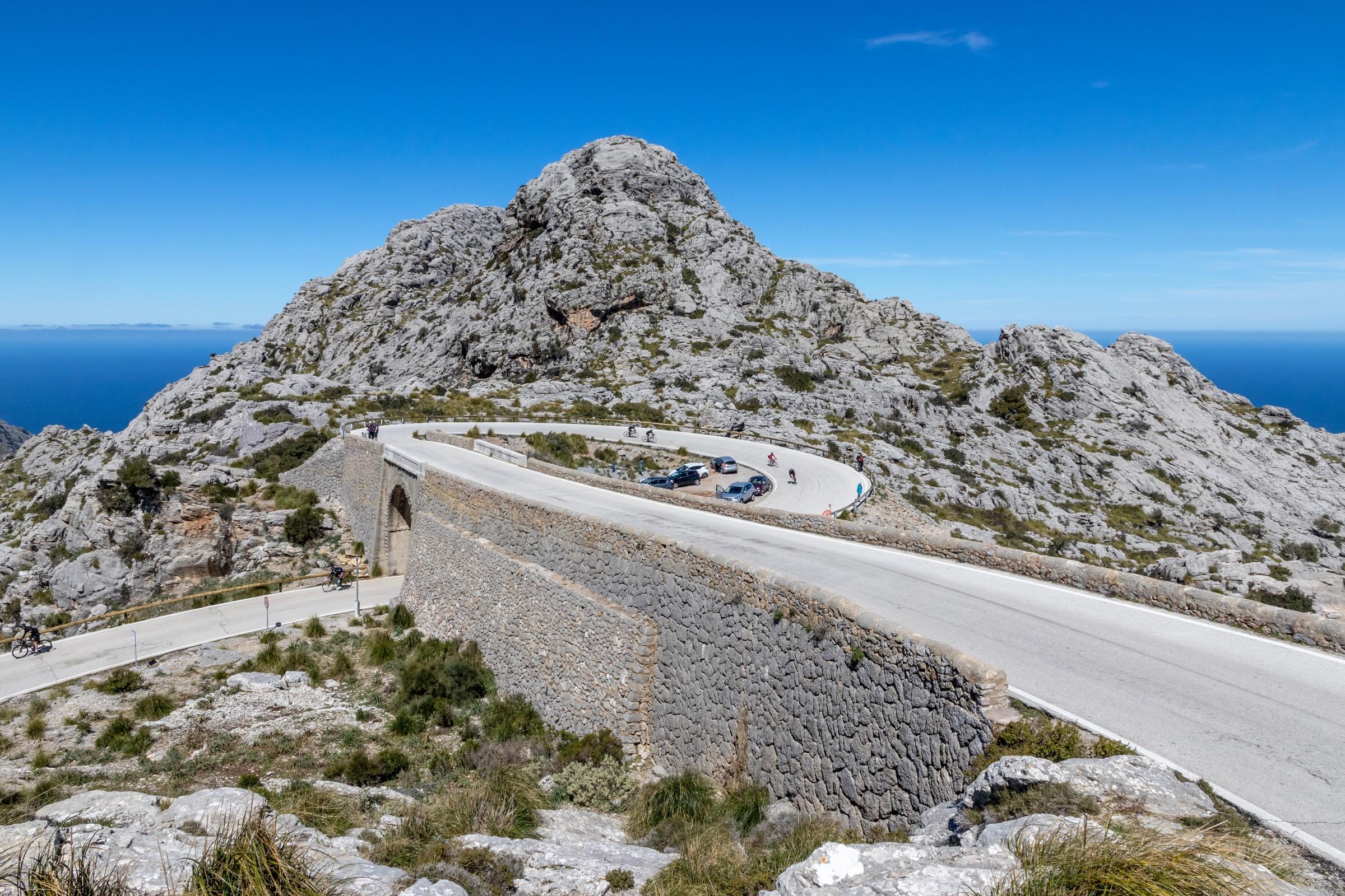 The Nus de sa Corbata loops around a rocky slope, with cyclists on the lower road passing beneath its stone bridge