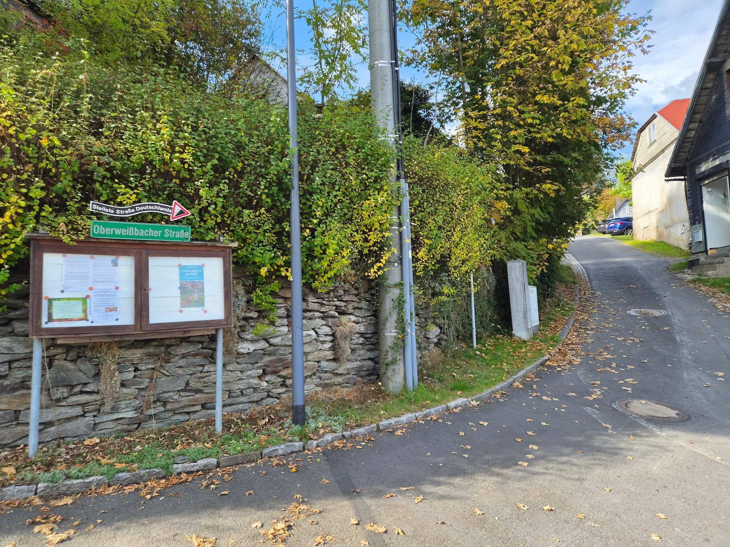 Oberweißbacher Straße rising beside houses and a stone wall, with its street-name sign and a noticeboard at the foot