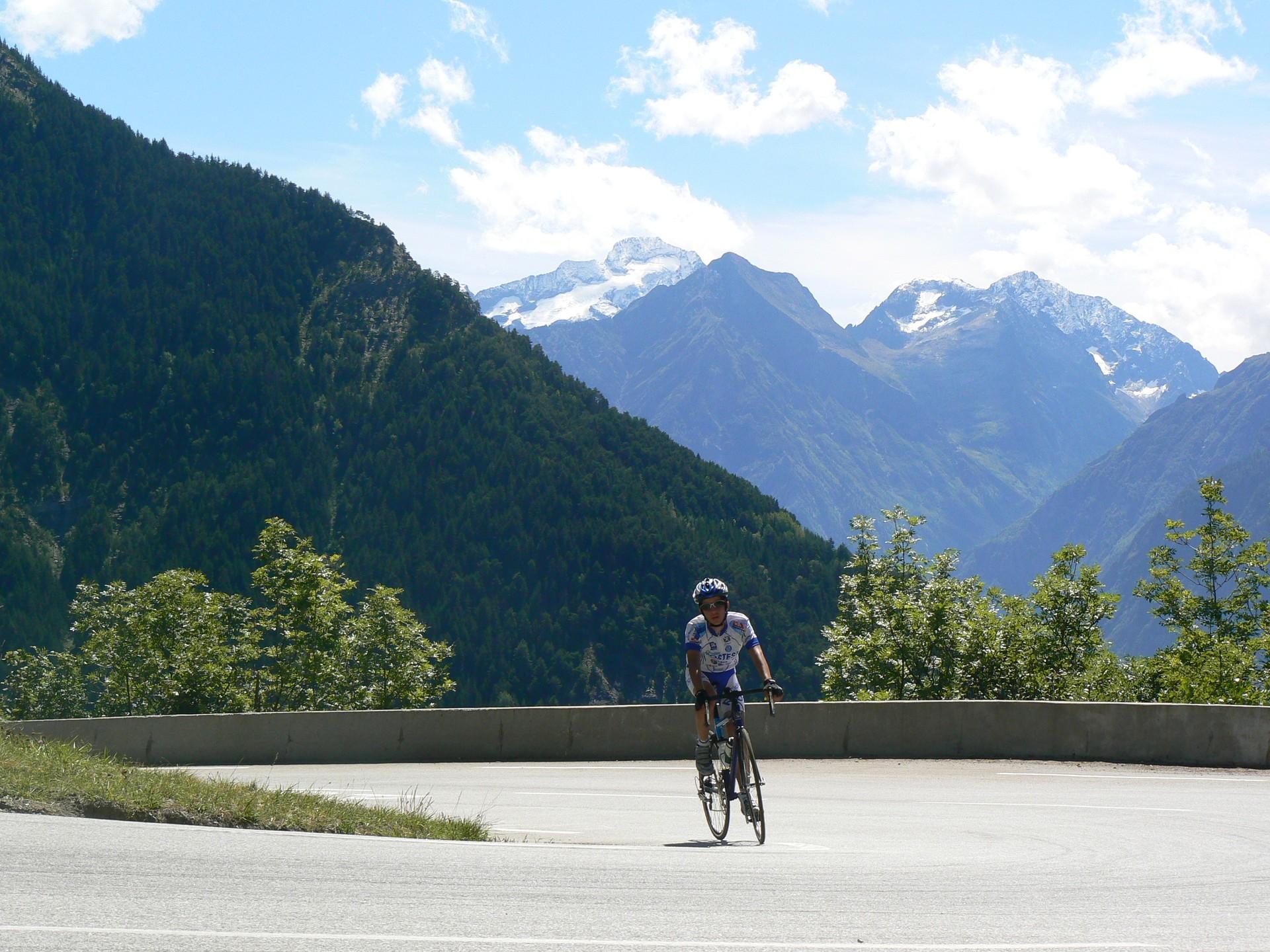 A cyclist rounding a bend on Alpe d’Huez, with wooded slopes and mountains behind
